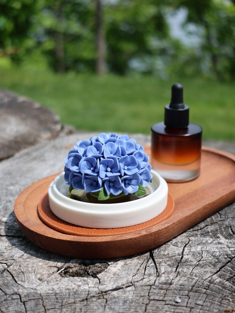 A vibrant blue hand-pinched porcelain hydrangea diffuser on a rustic wooden tray, set against a lush green natural background.