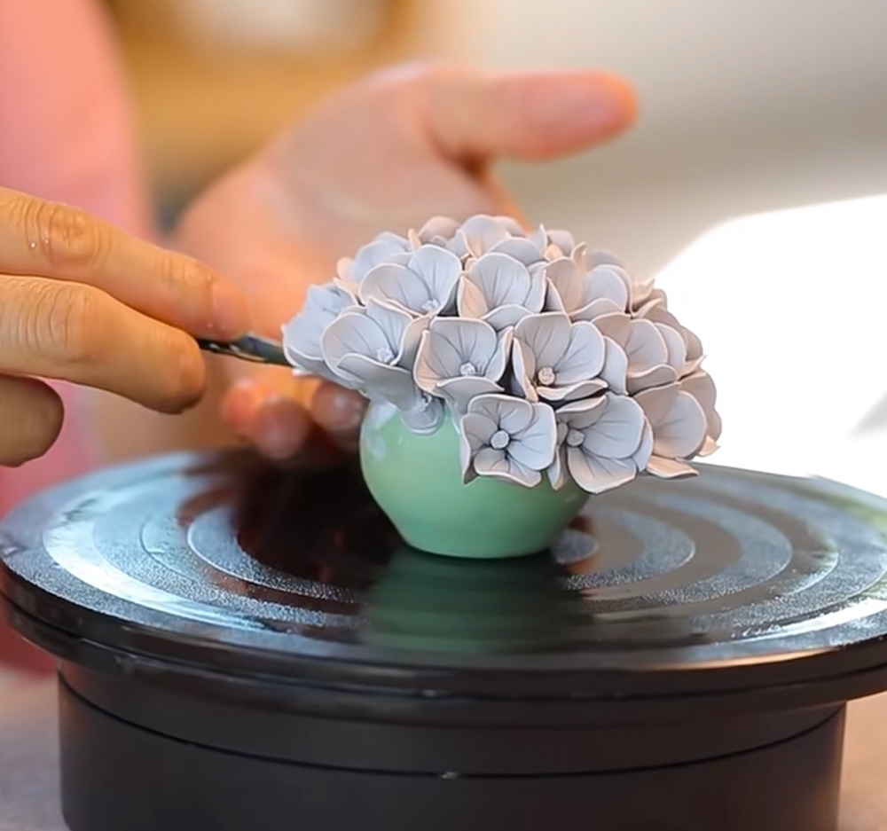 An artisan meticulously crafting a porcelain hydrangea bloom on a turntable, showcasing the delicate hand-pinching process of Home Is Home.